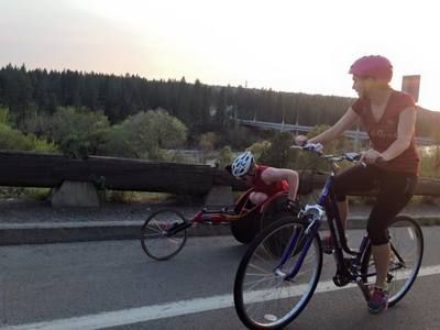 Cyclist in a pink helmet rides beside a racer along a riverside road