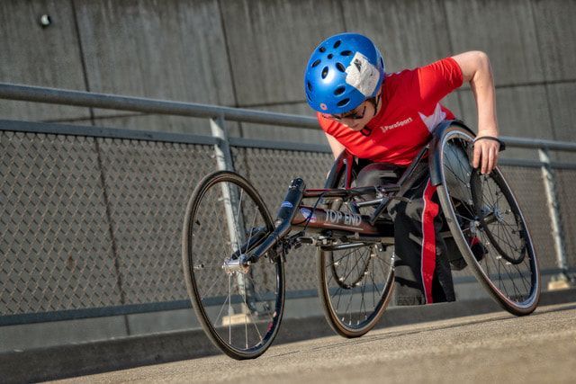 Young racer in a blue helmet and red ParaSport shirt pushes across a bridge