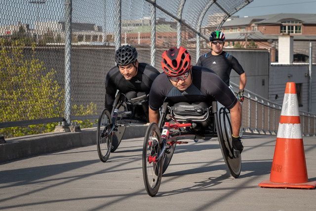 Three racers in black, one in a red helmet, pass an orange cone on a bridge