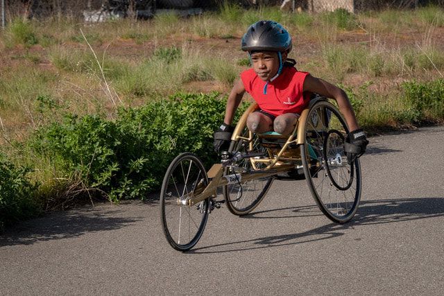Young athlete in a red shirt pushes a gold racing wheelchair past tall grass