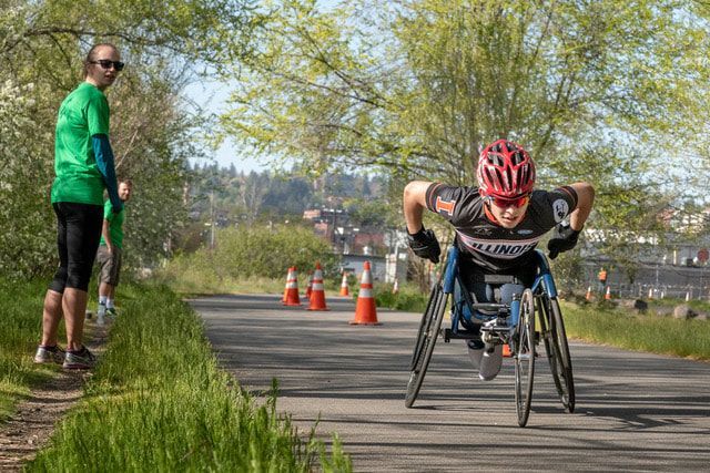 Racer in a red helmet and Illinois jersey heads up the trail past a volunteer