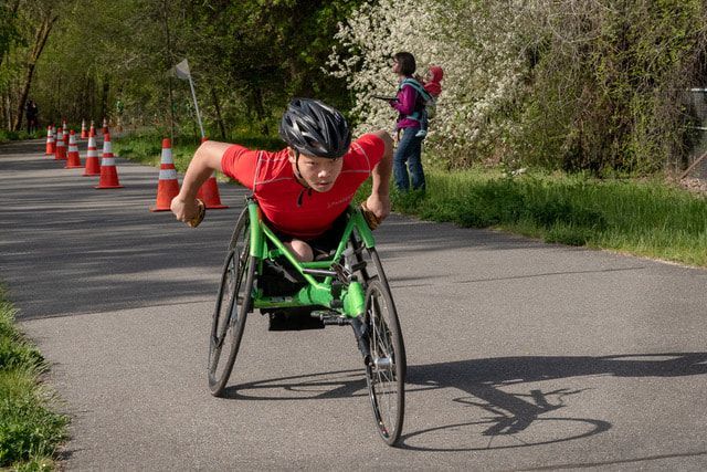 Racer in a red shirt drives a green racing wheelchair up the path past cones