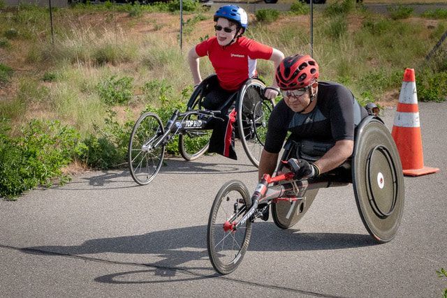 Racer in a red helmet leads a teammate in a blue helmet past an orange cone