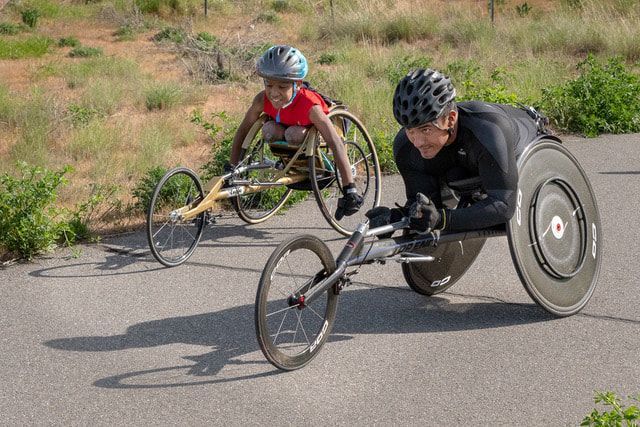 Racer in black leads a young racer in a gold chair along a weedy path