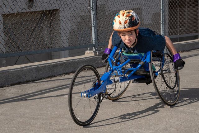 Young racer in a patterned helmet pushes a blue racing wheelchair by a fence