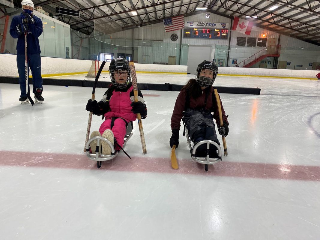 Two young sled hockey players hold sticks on the ice as a coach watches