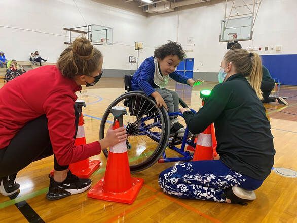 Two masked volunteers kneel by cones as a child in a wheelchair rolls past