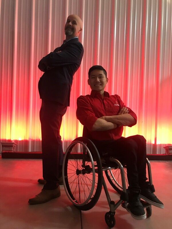 Two men pose back to back, one standing and one in a wheelchair