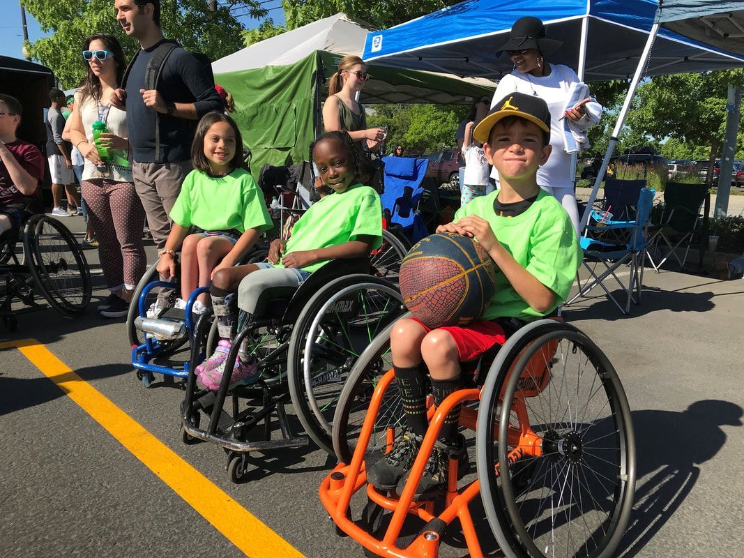 Kids in green shirts sit in sport wheelchairs at Hoopfest, one holds a ball