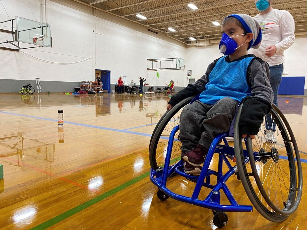 Masked young child sits in a blue sport wheelchair on a gym court