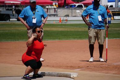 Shot putter in red crouches to throw as officials in blue watch