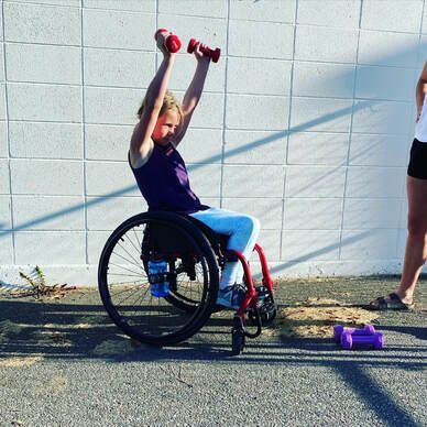 Young athlete in a wheelchair lifts red dumbbells overhead outdoors