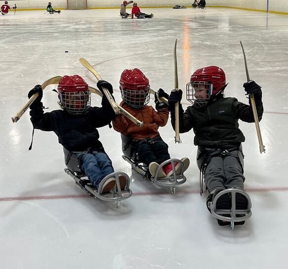 Three young sled hockey players in red helmets raise their sticks on the ice