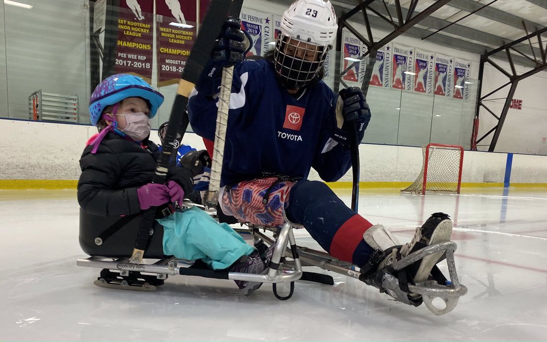 Child in a blue helmet and player #23 sit in their sleds near the hockey net