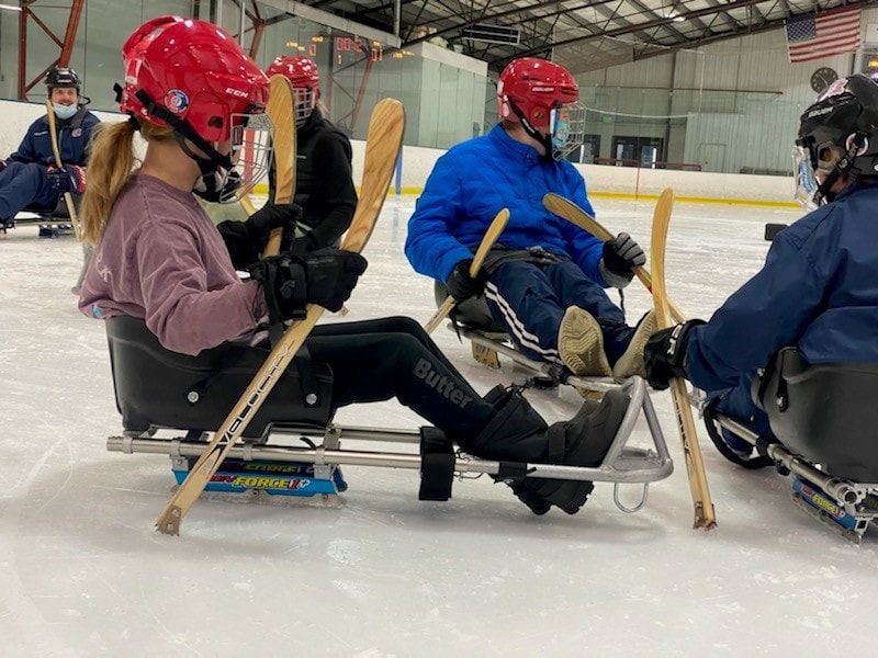 Sled hockey players in red helmets line up with their sticks on the ice