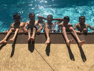 Five young swimmers sit on the pool edge with the water behind them