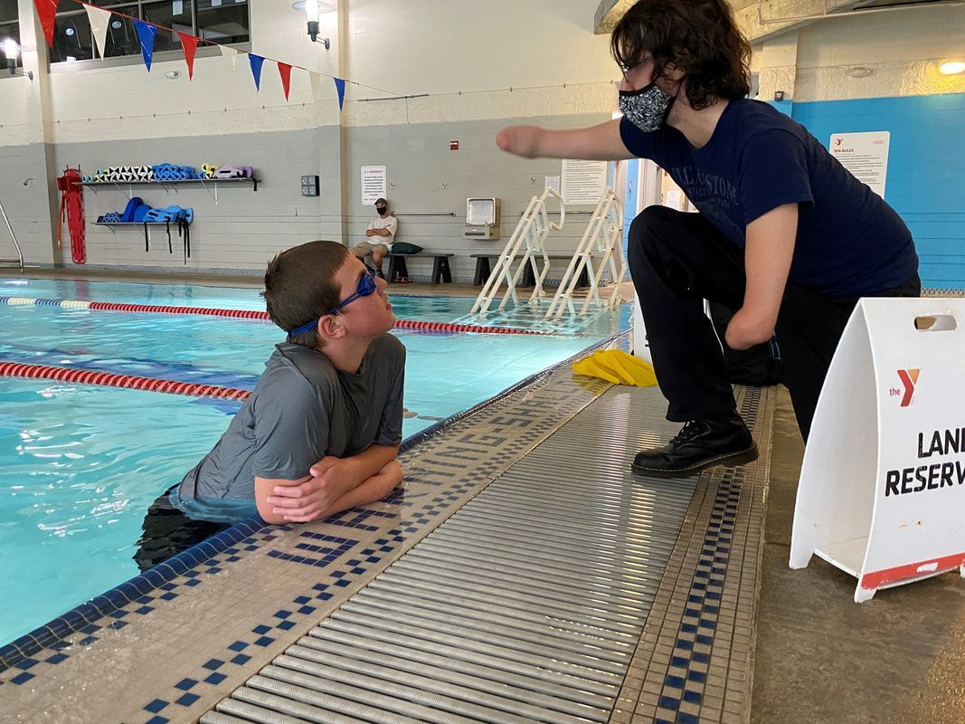Swim coach kneels at the YMCA pool edge talking with a swimmer in goggles