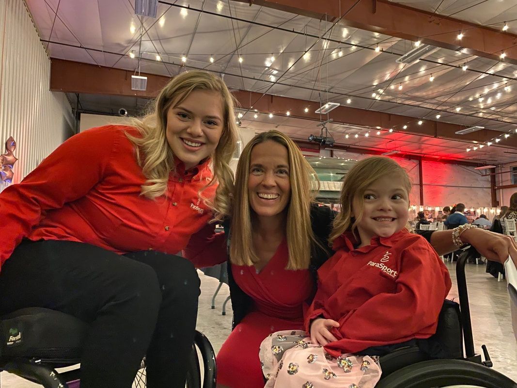 Two athletes in wheelchairs and a woman in red smile together at an event