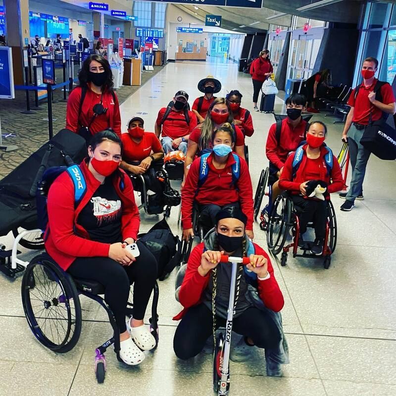 Masked team in red poses in an airport terminal, one athlete with a scooter
