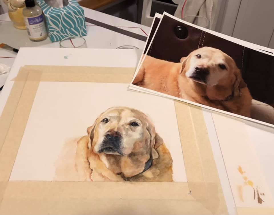 A dog portrait painting with a reference photo of a smiling yellow Labrador on a desk.
