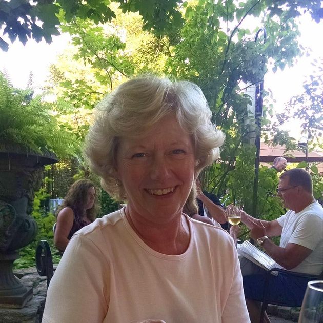 Smiling woman in a white shirt seated outdoors at a garden café, with people dining behind her.