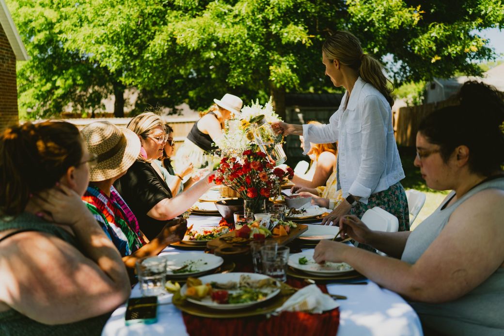People smiling, drinking wine, and eating at a table, likely friends or family, in a brightly lit kitchen.