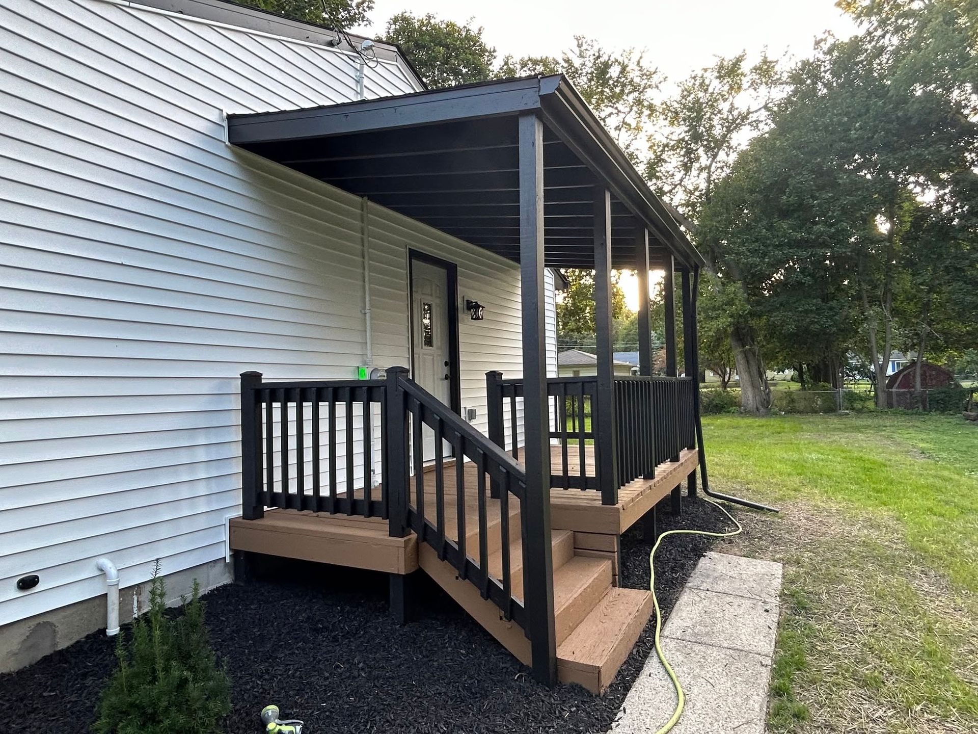 Small covered wooden porch with steps and black railings beside a white house