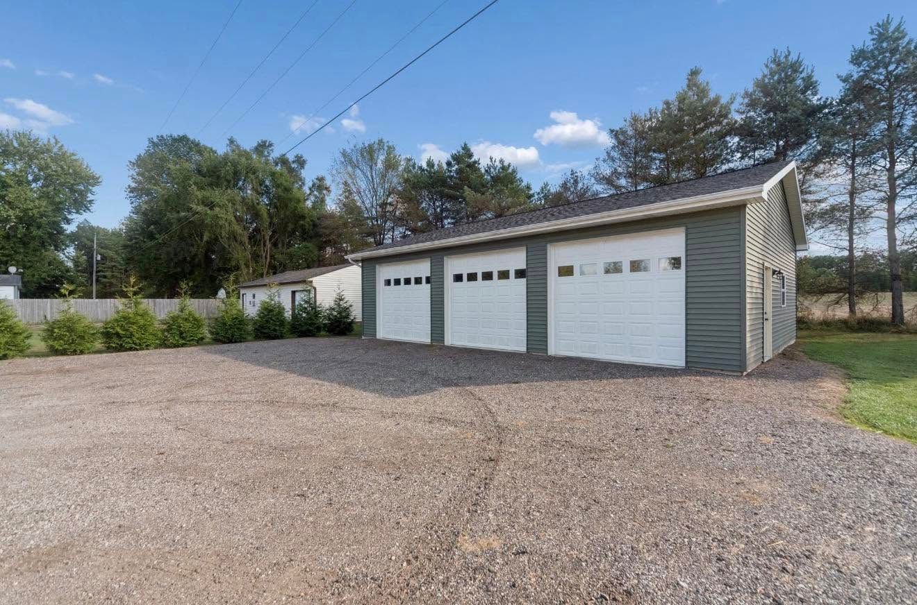 Gravel driveway leading to a white three-car garage with green siding and trees under a blue sky