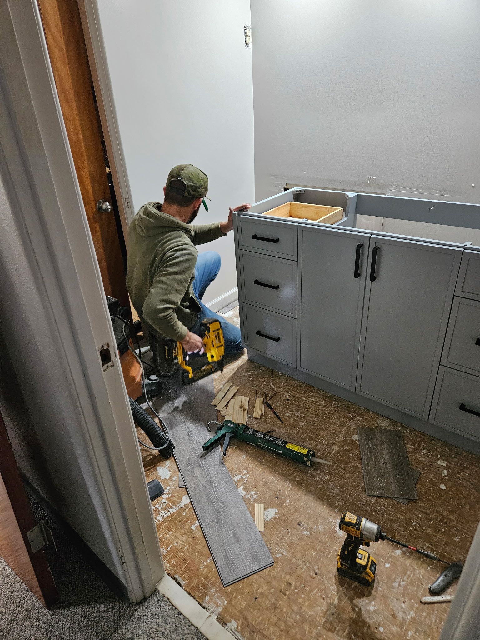 Worker kneeling in a narrow utility room, installing a large gray appliance with tools and debris on the floor