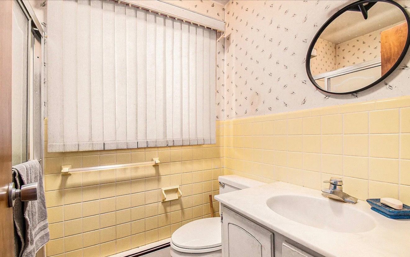 Small yellow-tiled bathroom with sink, toilet, oval mirror, and closed blinds over the window.