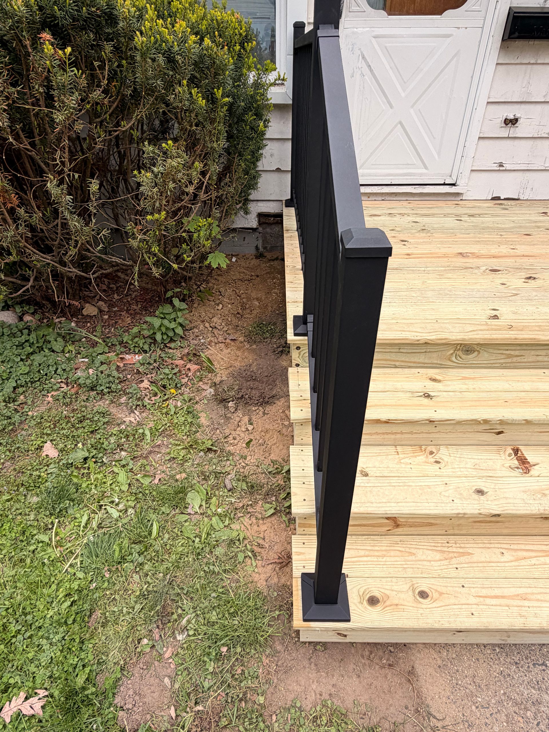 Wooden porch stairs with a black railing beside a grassy yard and bush.