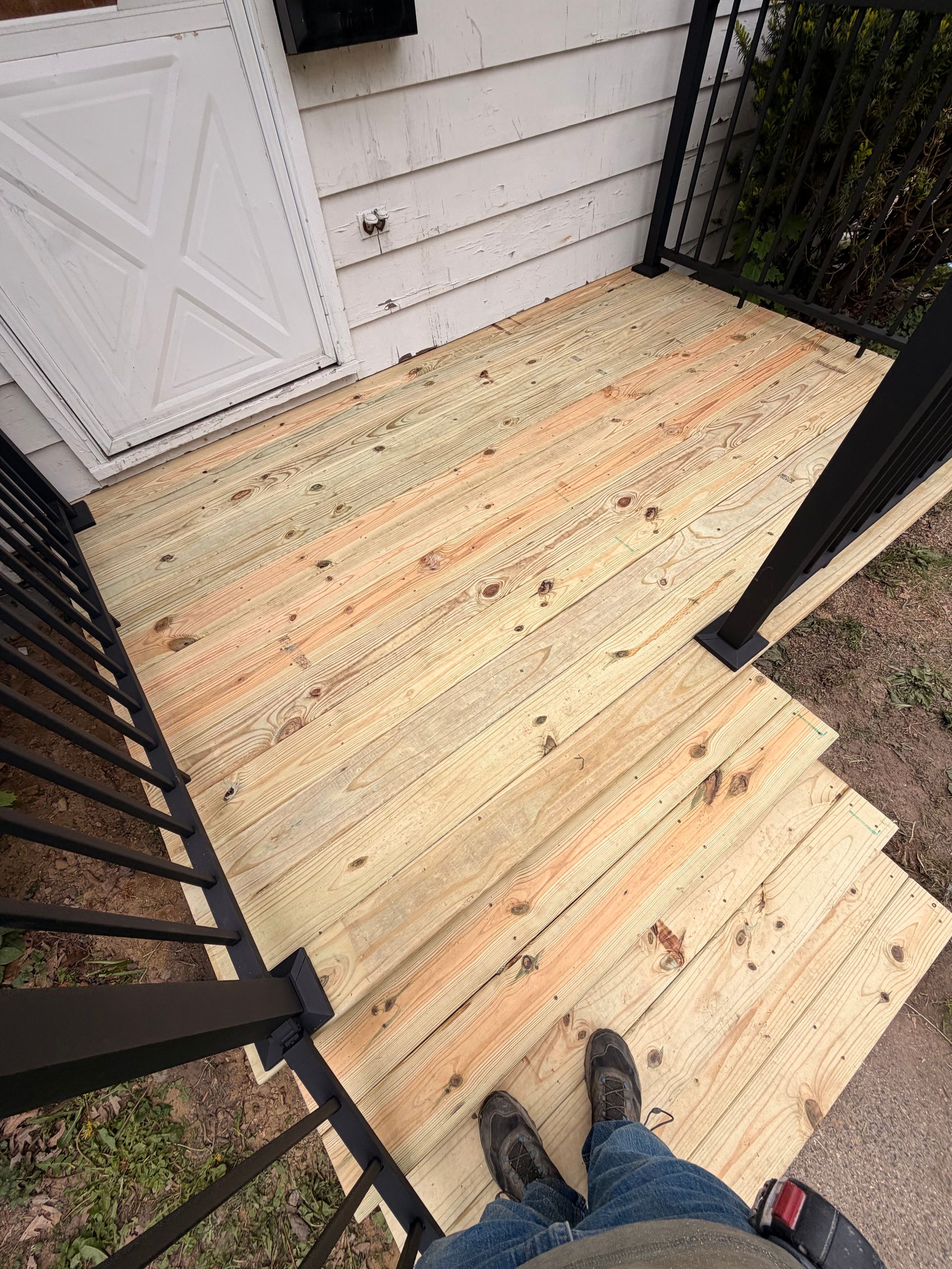 Wooden porch steps and landing beside a white building, viewed from above with a person’s feet visible.