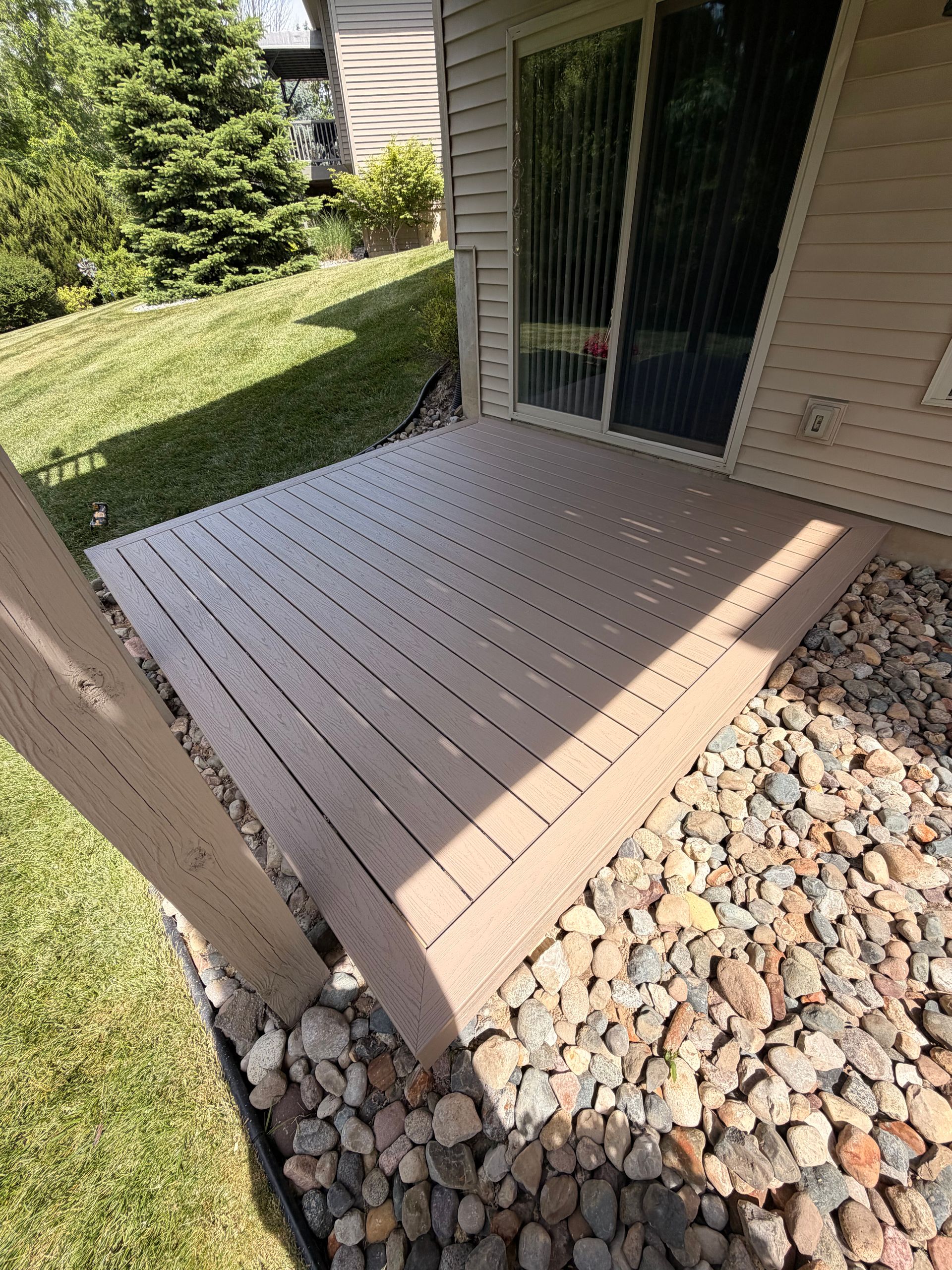Small tan wooden patio deck beside a sliding glass door, bordered by grass and river rocks.