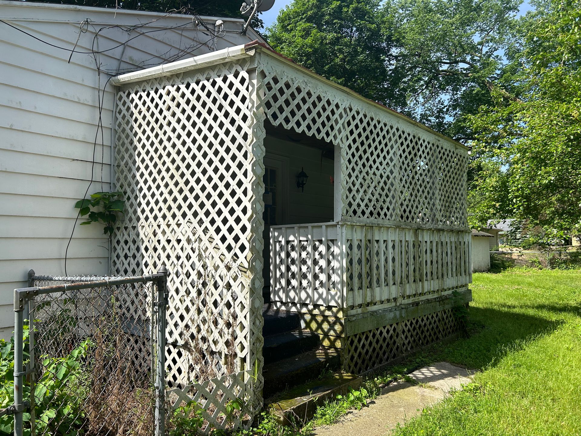 White house side porch with white lattice railing and steps, beside a grassy yard and trees