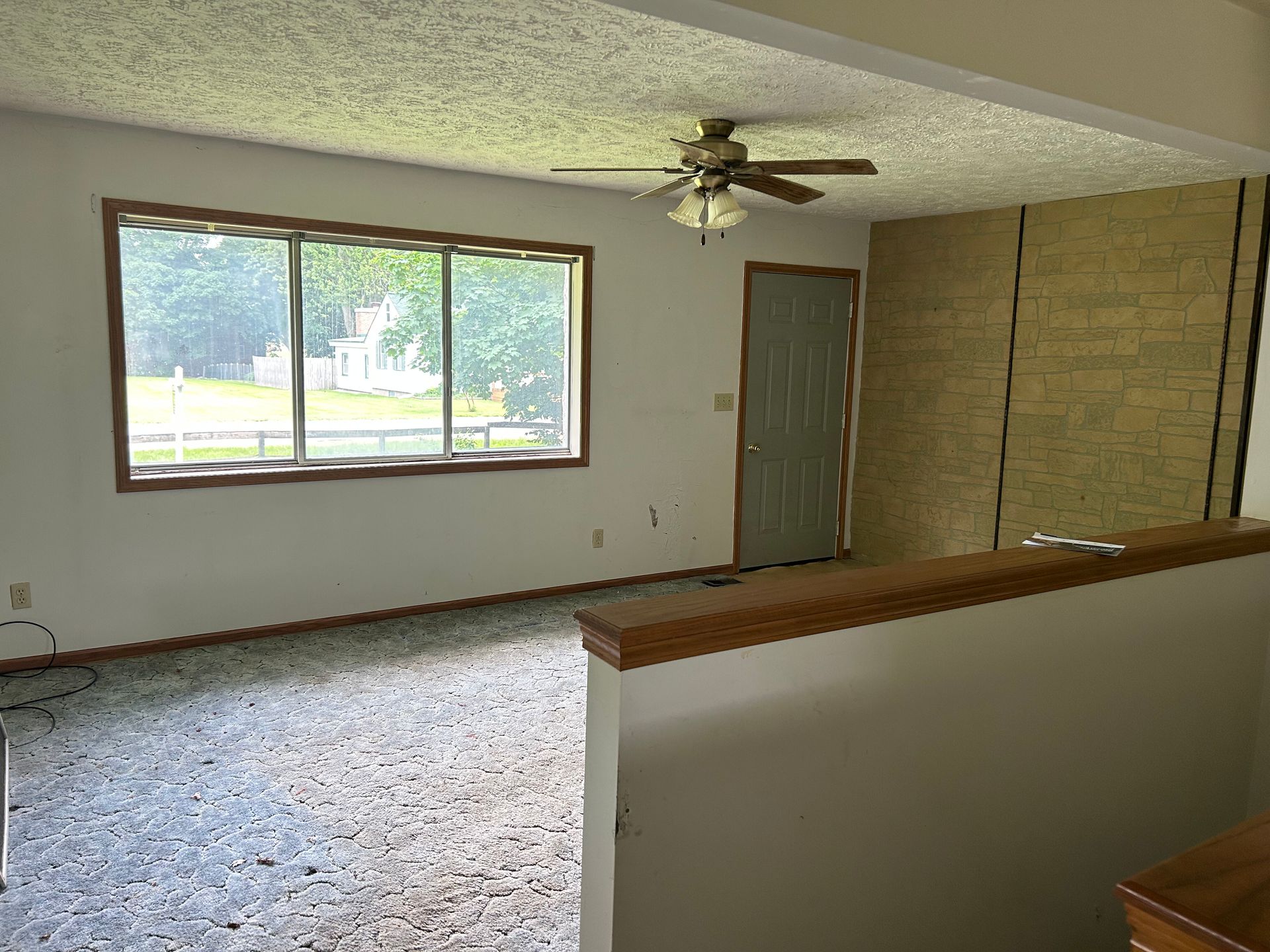 Empty living room with carpet, large window, ceiling fan, and wood-paneled wall.