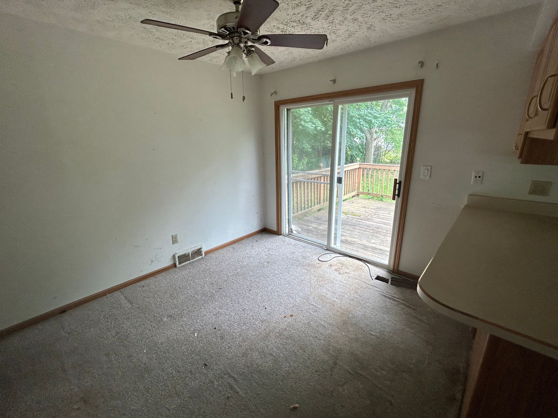 Empty carpeted room with ceiling fan, white walls, and sliding glass door to a deck.