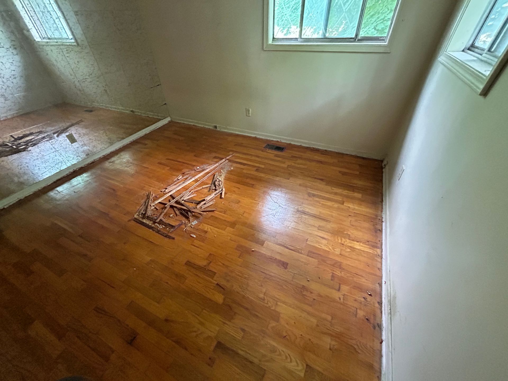Empty hardwood room with bare walls, windows, and a small pile of debris on the floor