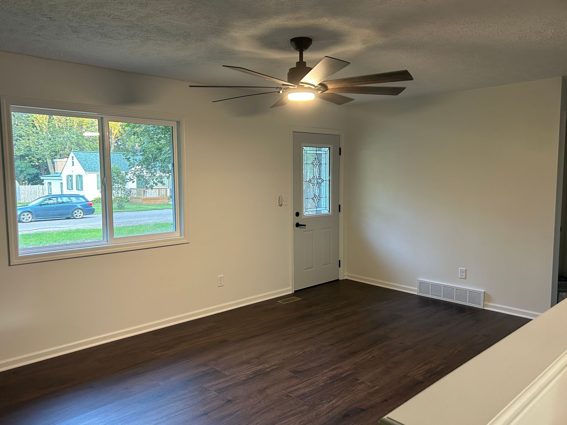 Empty living room with dark wood floors, ceiling fan, window, and front door.