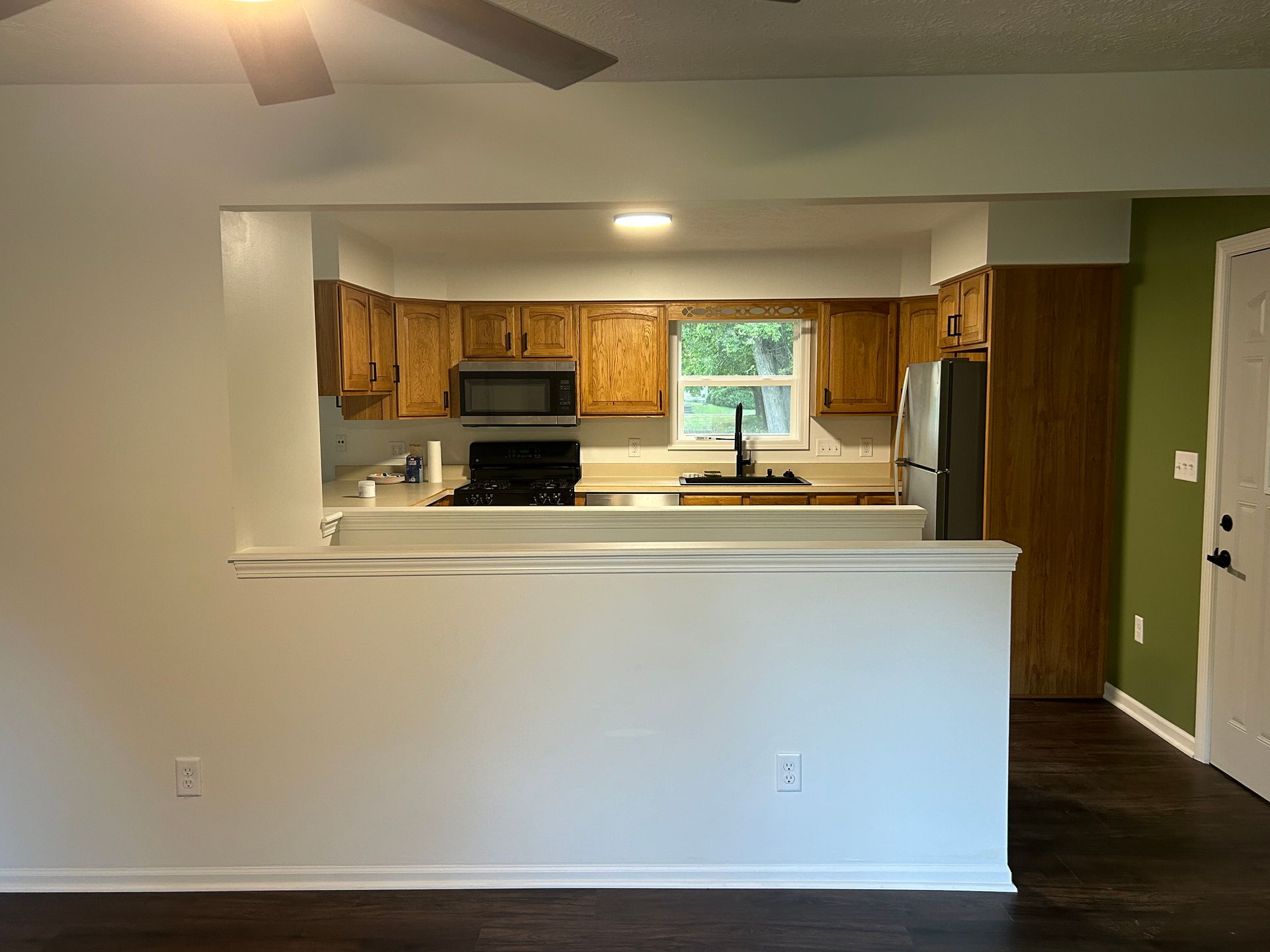 Open kitchen viewed from a living room, with wood cabinets, white breakfast bar, and green accent wall.