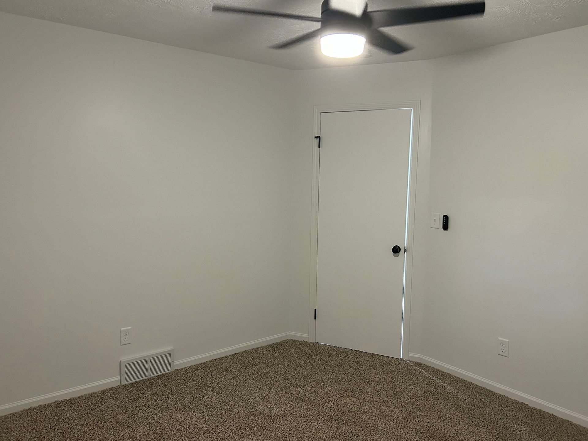 Empty carpeted room with white walls, a ceiling fan, and a closed door.