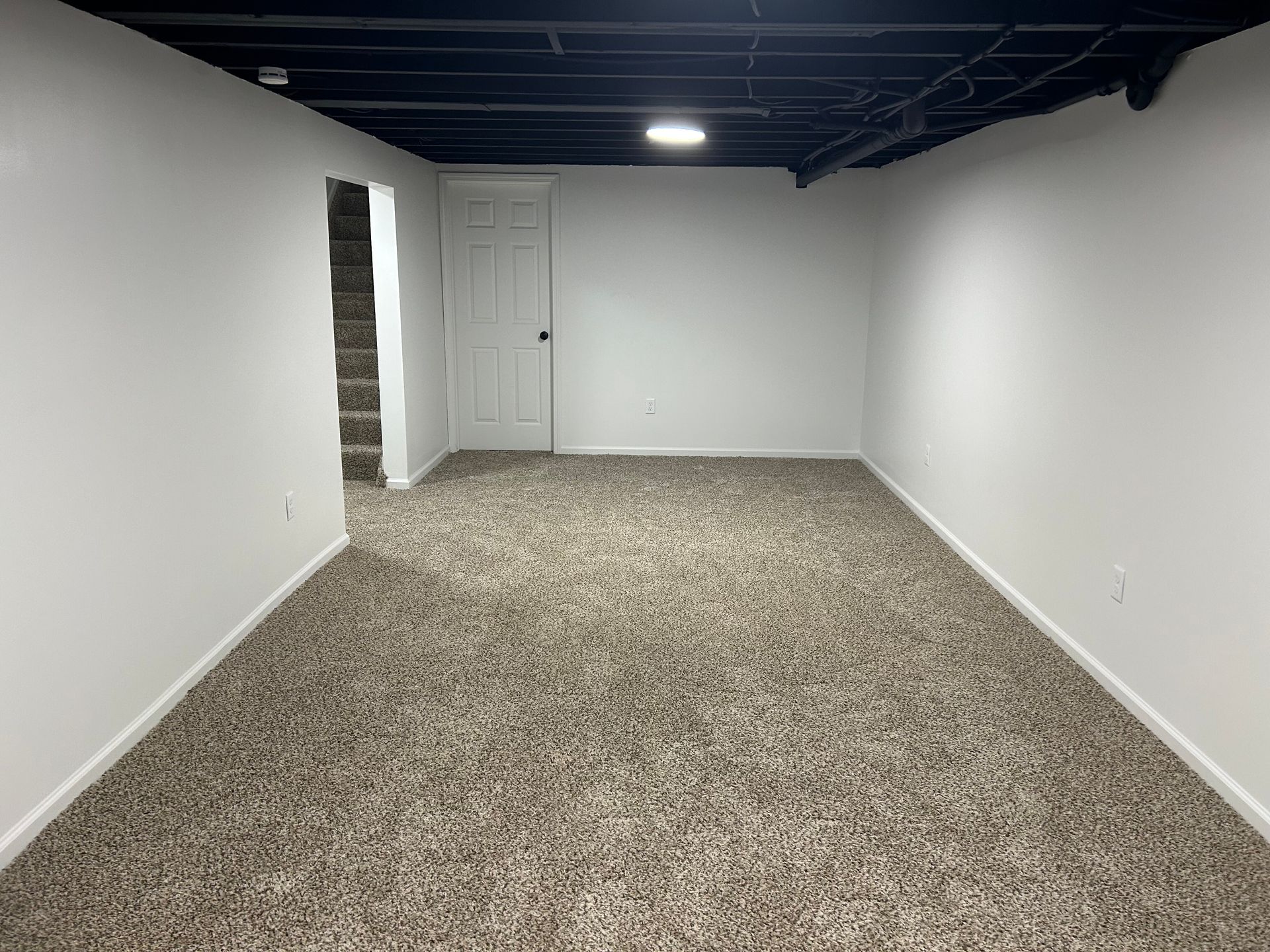 Empty basement room with white walls, carpeted floor, and exposed black ceiling beams.