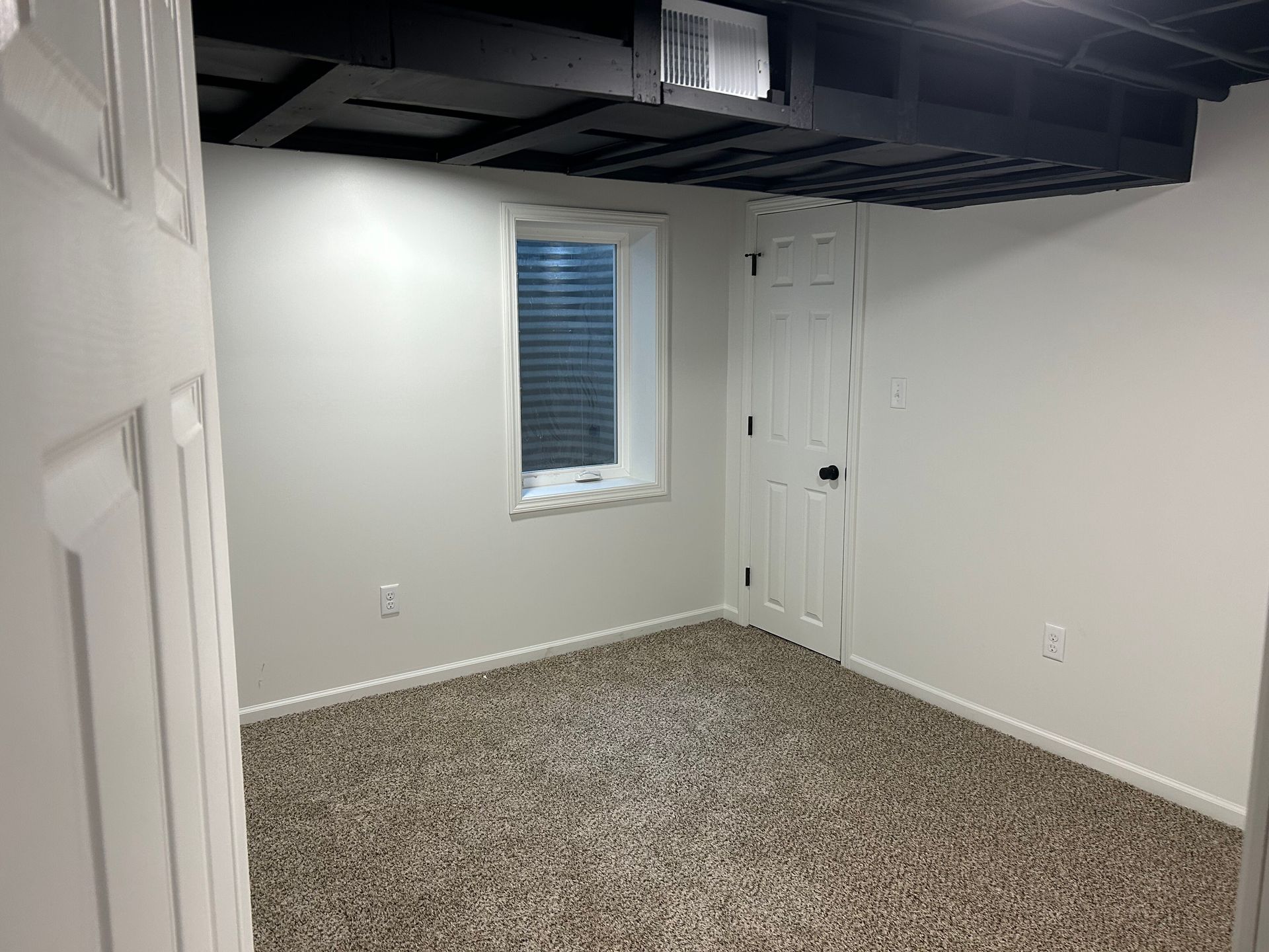 Empty basement room with white walls, speckled carpet, small window, and exposed black ceiling.