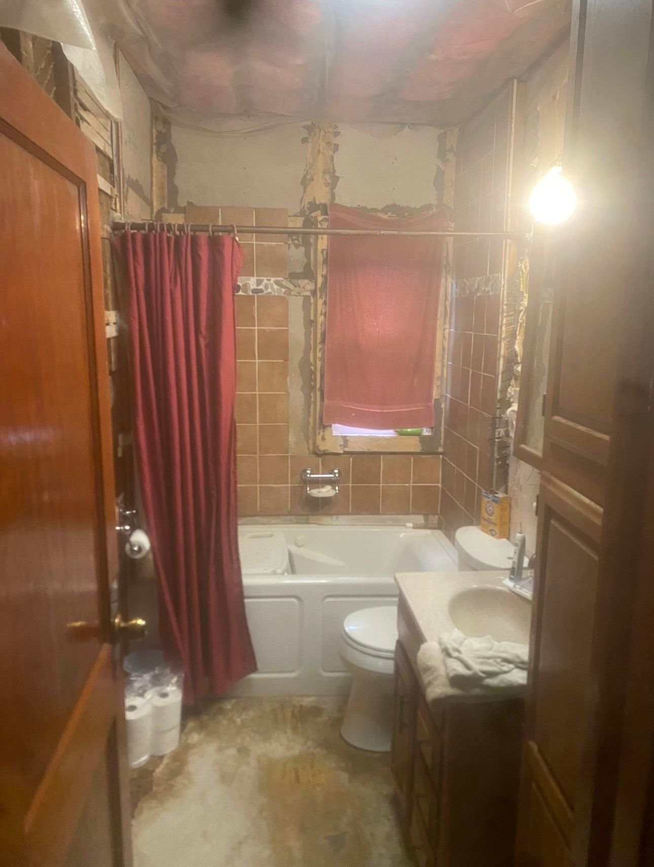 Small bathroom with red shower curtain, white tub and toilet, and a sink under a bright light.