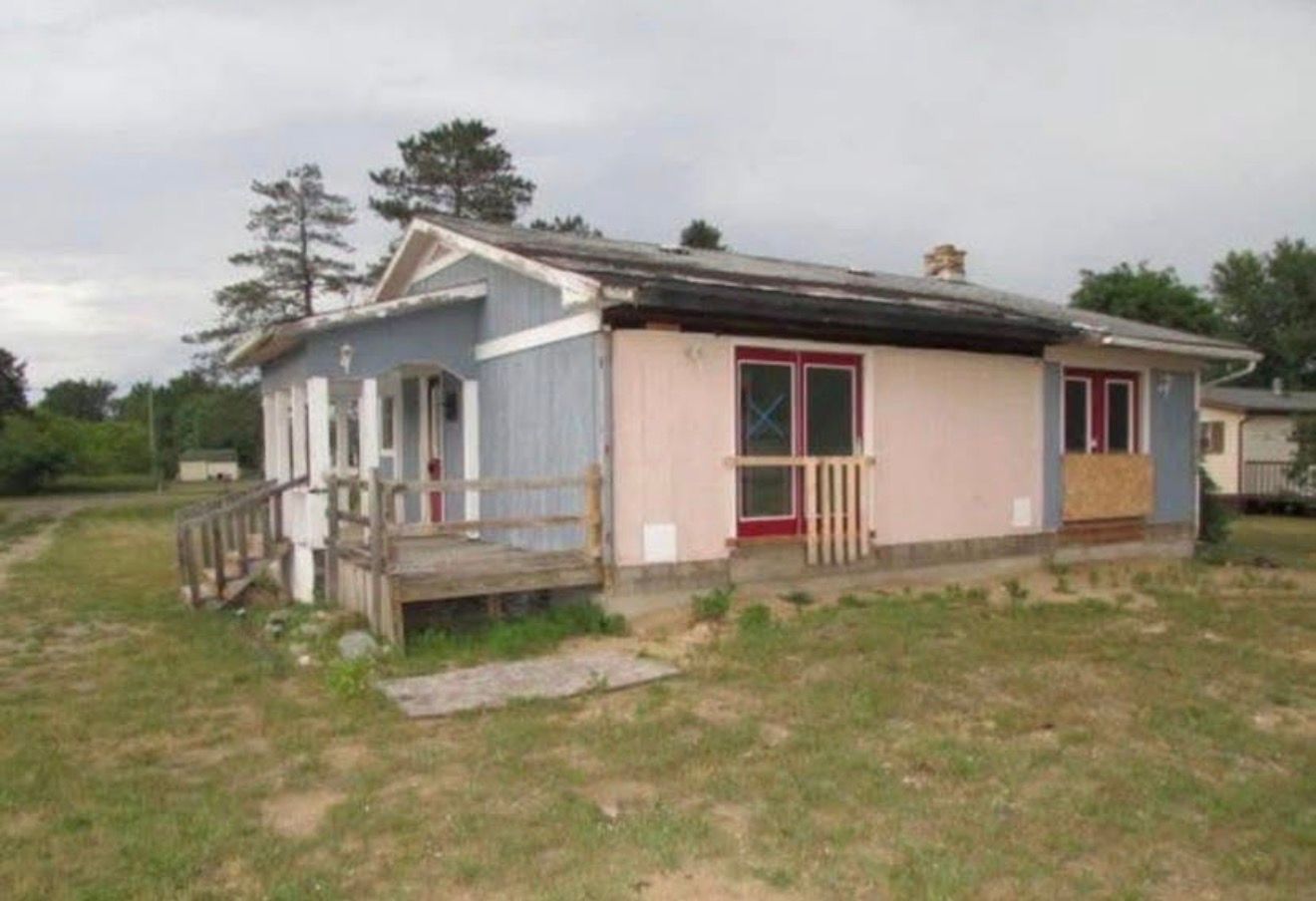Small weathered rural house with a porch, pink siding, and a grassy yard under an overcast sky
