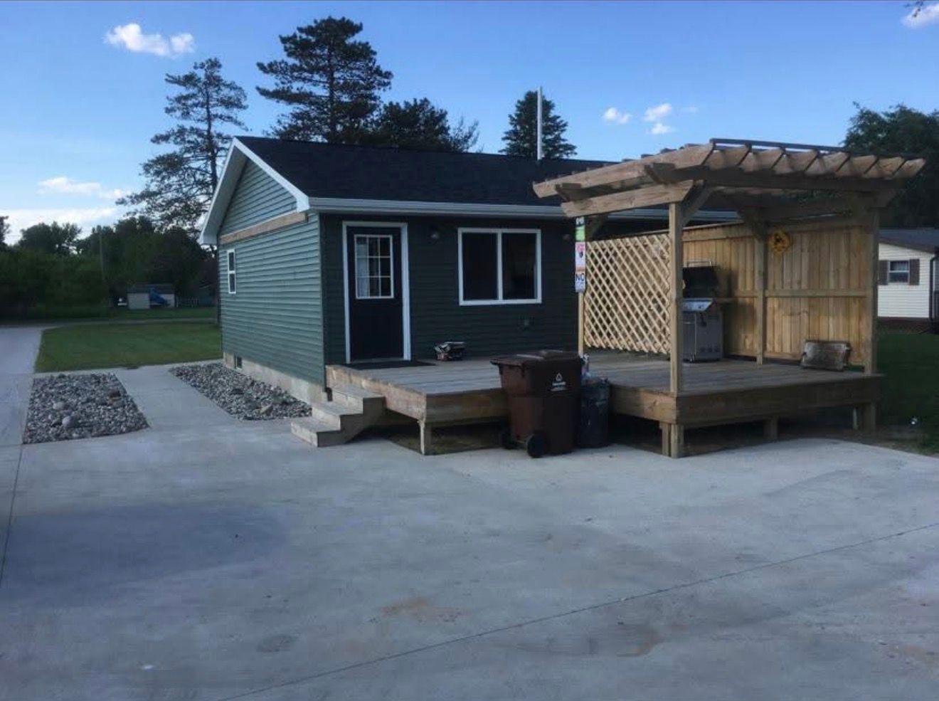 Small dark cabin with a wooden deck and pergola on a paved lot at dusk