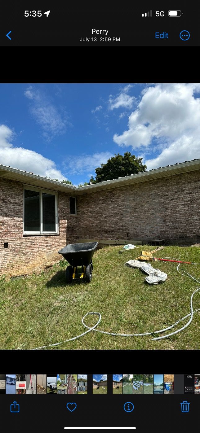 Backyard with a brick house wall, wheelbarrow, lawn, and cloudy blue sky