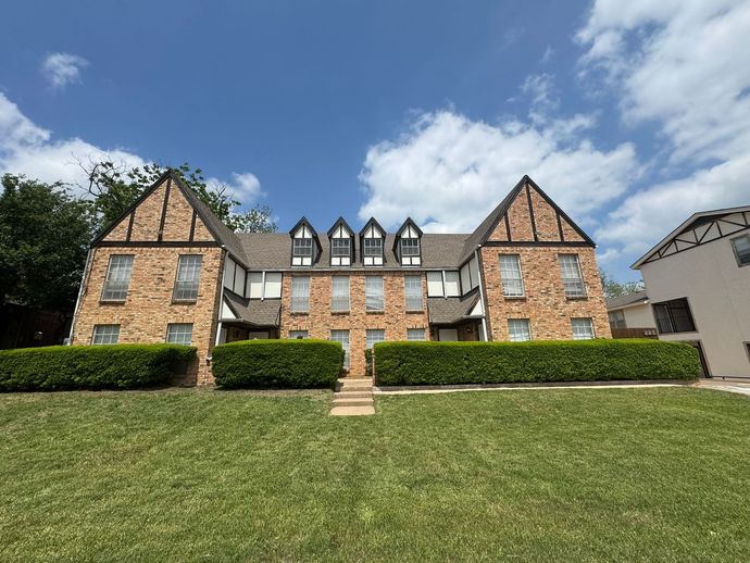Stone apartment building with steep gables, hedges, and a grassy lawn under a partly cloudy sky