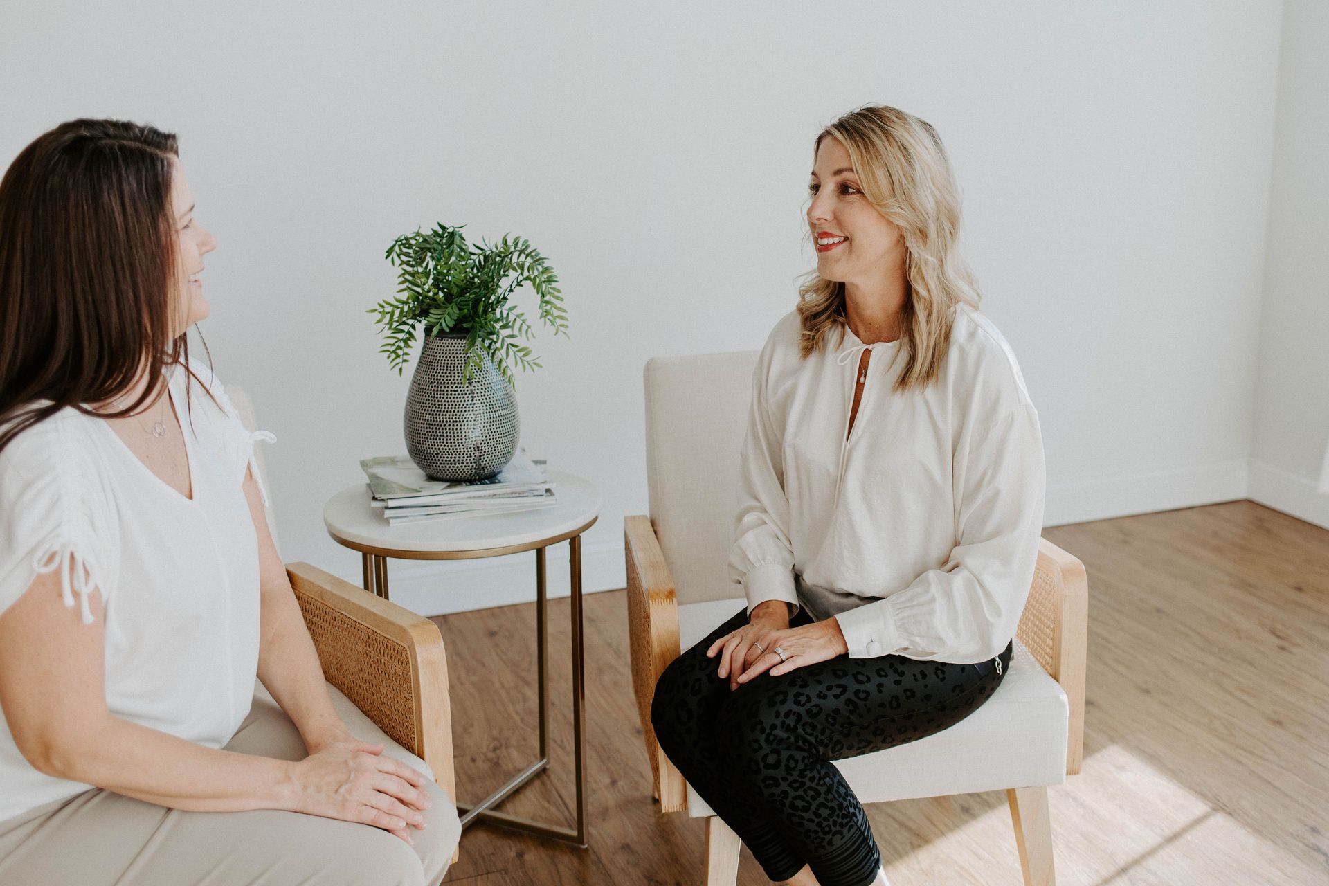 Two women sit facing each other in a bright room, discussing something, a plant on a small table.