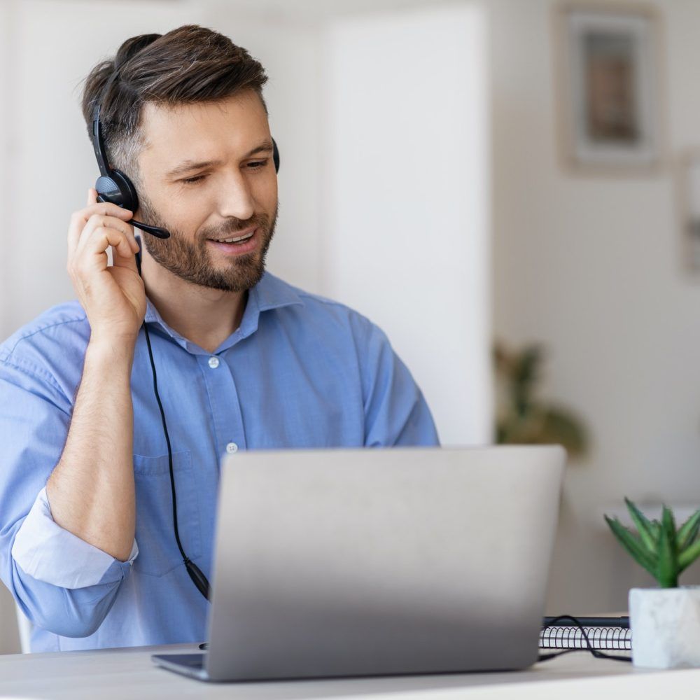 Man wearing headset, using laptop, smiling. Indoors, bright.