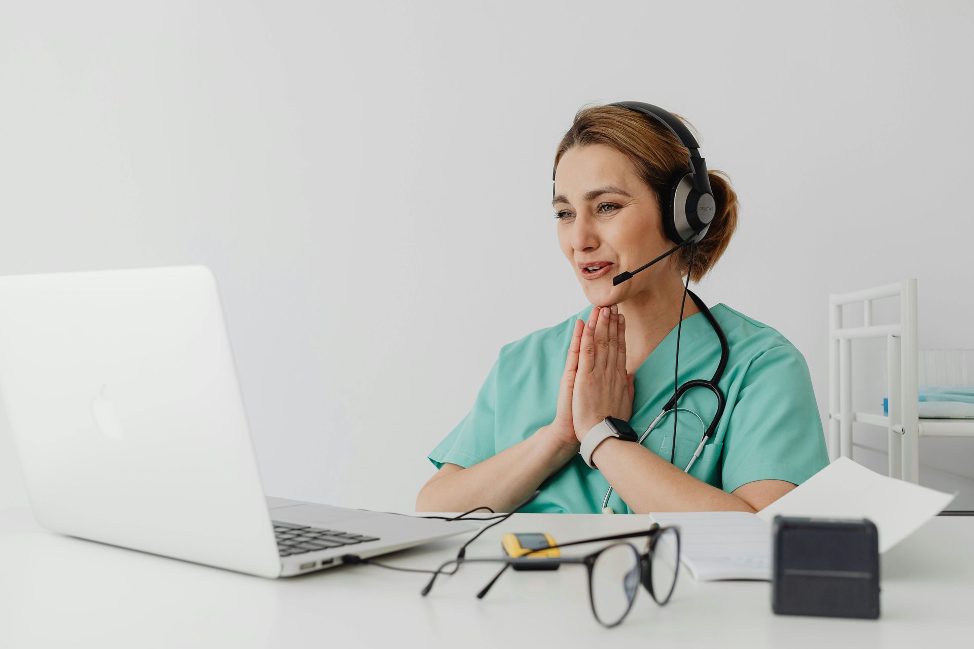 Healthcare worker in scrubs on a video call, wearing a headset, hands clasped, laptop open, stethoscope.